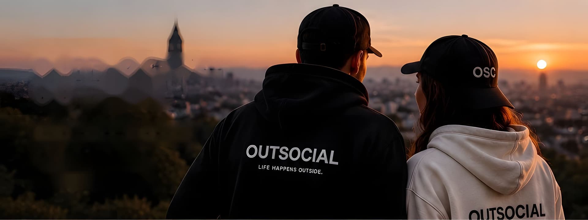 Two friends seen from behind at sunset over a city, in an OUTSOCIAL hoodie and OSC caps.