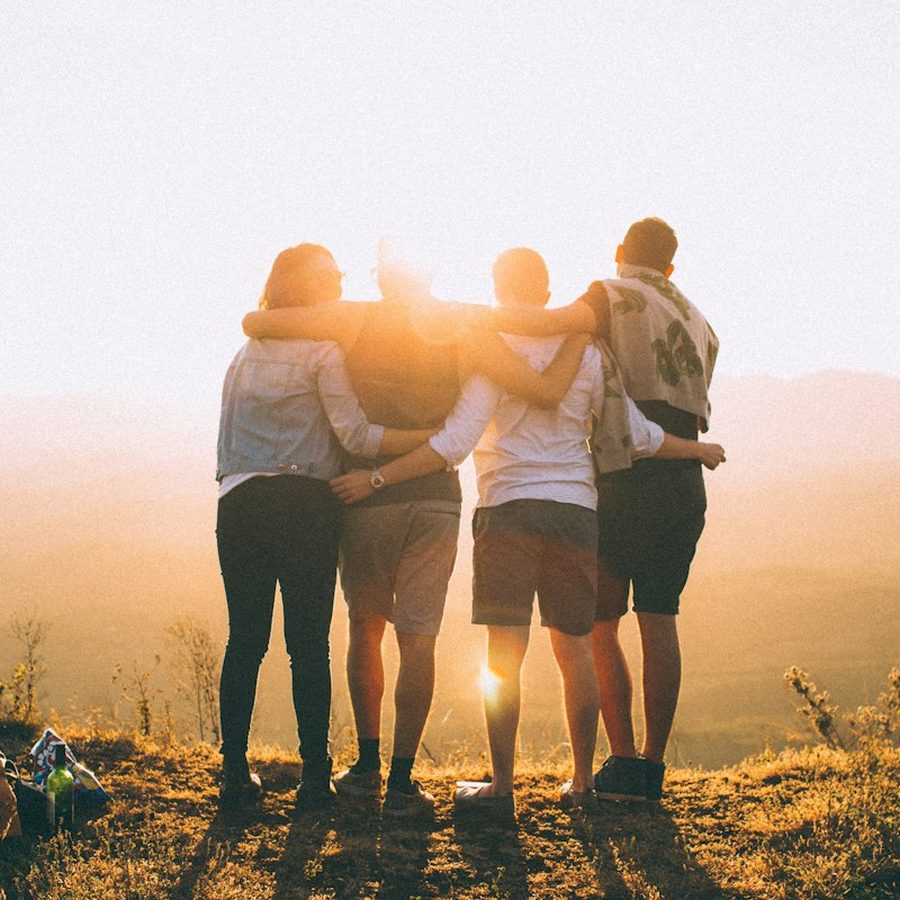 Four friends with their arms around each other watching the sun set from a hill.