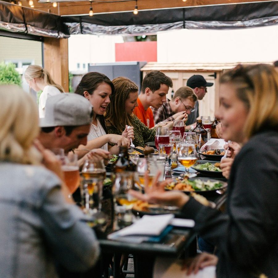 A big group sharing food and drinks around one long table on a terrace.